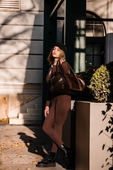 Fashionable young brunette woman with long legs. wearing Solid Long Sleeve Bodycon One Piece Jumpsuits posing with a leather brown travel bag on city street in old town