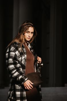 Outdoor fashion portrait of young beautiful fashionable girl wearing trendy checkered shirt, solid long sleeve bodycon one piece jumpsuits and small brown color small bag, posing in street.