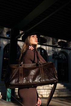 Fashionable young brunette woman wearing Solid Long Sleeve Bodycon One Piece Jumpsuits posing with a leather brown travel bag on city street in old town