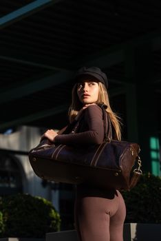 Fashionable young brunette woman wearing Solid Long Sleeve Bodycon One Piece Jumpsuits posing with a leather brown travel bag on city street in old town