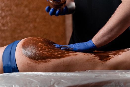 The masseur smears chocolate on the legs and thighs of the girl in the spa center. Beauty procedure chocolate wrap