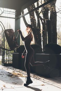 Fashionable young brunette woman with long legs. wearing solid long sleeve bodycon one piece jumpsuits posing with a leather brown travel bag on city street in old town