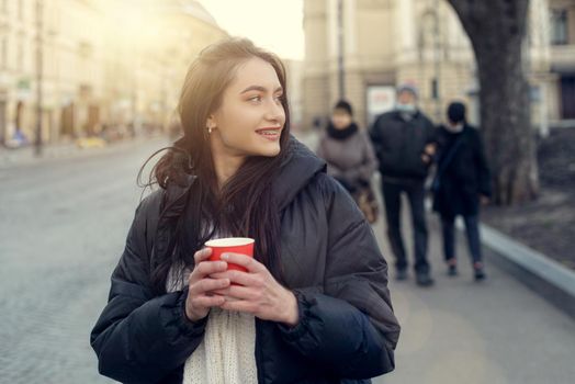 Stylish Caucasian woman in a coat stands and drinks coffee on the street. Urban style and fashionable girl with a paper cup of coffee.