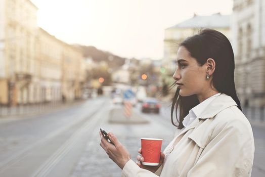 A woman on the street uses a mobile phone. online shopping. use of mobile applications. beautiful woman with long dark hair in a raincoat