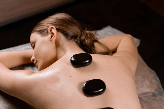 Hot stone massage therapy for relax and ease tense muscles and damaged soft tissues of body. Heated stones are placed on specific parts young woman.