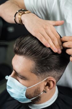 Hair styling with wax for a bearded man in a medical mask. The work of the barbershop in quarantine coronavirus covid-19.