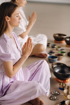 Two women are sitting with Tibetan bowls in the lotus position before a yoga class in the gym.