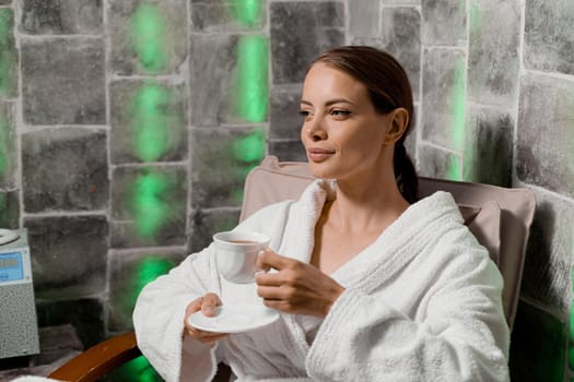 Attractive woman in bathrobe with cup of tea relaxing in salt room in spa
