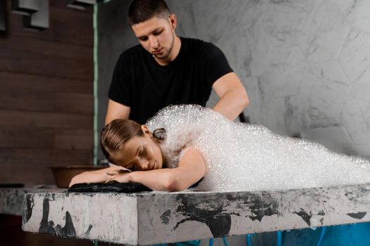 Foam peeling in Turkish spa hammam. Attractive model is relaxing in spa. Masseur is making massage using sponge glove