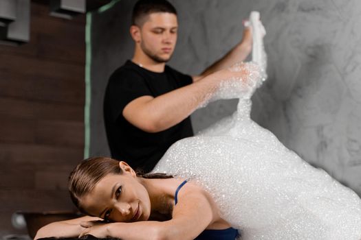 Attractive girl relaxing in spa. Masseur is making foam peeling in Turkish spa hammam