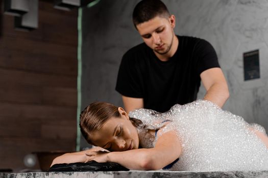 Foam peeling in Turkish spa hammam. Attractive model is relaxing in spa. Masseur is making massage using sponge glove