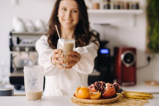 A happy smiling healthy woman has prepared a fruit cocktail and gives it to you while standing at home in the kitchen.Healthy eating.