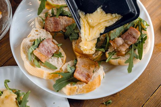 Raclette melted cheese for sandwich with fried bacon and arugula traditional french cuisine. Plate with snacks
