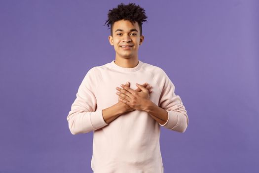 Portrait of romantic handsome hispanic man with dreads, hold hands pressed to heart and smiling pleased, see heartwarming nice gesture, receive lovely gift, being touched, purple background.