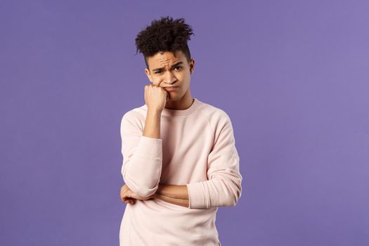 Portrait of complicated, troubled young hispanic man facing tough problem, lean on fist grimacing and pouting, solving troublesome situation, thinking, feeling uneasy, purple background.