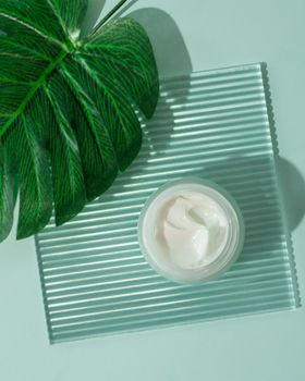 Cosmetic cream or moustirizer on transparent ribbed acrylic plate with tropical palm monstera leaf over blue background. Open round jar, aesthetic swirls cream. Top view flatlay. Copy space. Vertical