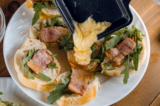 Raclette melted cheese for sandwich with fried bacon and arugula traditional french cuisine. Plate with snacks