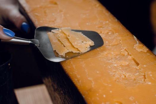 Slicing aged cheese parmesan with crystals using slicer knife. Hard cheese with knife for appetizer on dark background