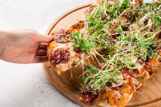 Piece of pinsa romana in hand with stretching mozzarella with salami, cheese, mushrooms, decorated with microgreens on wooden board