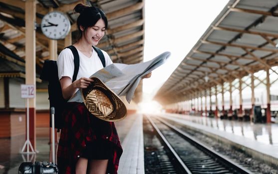 Young woman traveler with luggage and hat looking at map with train background at train station. travel concept