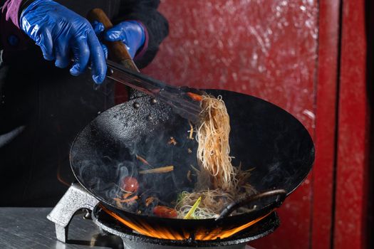 Funchoza rice noodles with vegetables cooking on fire in wok pan. Street flambe food.