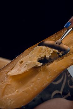 Slicing aged cheese parmesan with crystals using slicer knife. Hard cheese with knife for appetizer on dark background