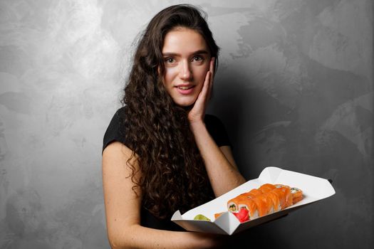 Surprised girl with sushi set philadelphia rolls in a paper box happy girl holding on a gray background. Food delivery