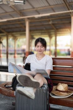 Happy young asian woman traveler or backpacker using map choose where to travel with luggage at train station, summer vacation travel concept.