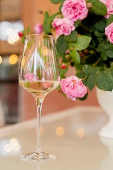 Glass of white wine against the background of bouquet roses in white vase on light table.