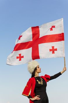 Georgian girl waving national flag of Georgia on blue sky background. Georgian culture lifestyle. Woman in papakha and red dress