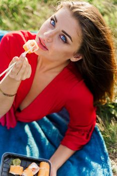 Sushi near lips of girl with blue eyes at picnic. Food delivery from japanese restaurant. Vertical photo advert for social networks. Pretty girl eats sushi set in the park.