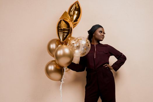 Black girl with golden ballons have a party and smile isolated on peach background. African woman celebrate graduation. Happy emotion of muslim young woman