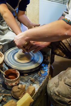 the Hands of a master and a student make a pitcher on a Potters wheel of yellow clay. Selective focus on hands