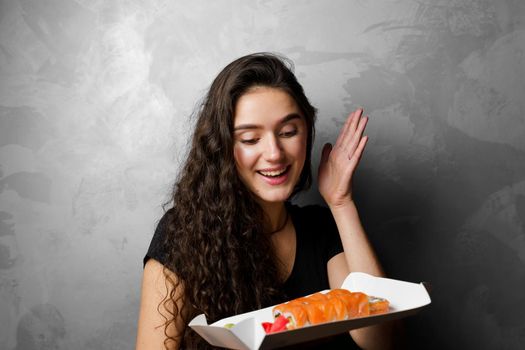 Surprised girl with sushi set philadelphia rolls in a paper box happy girl holding on a gray background. Food delivery