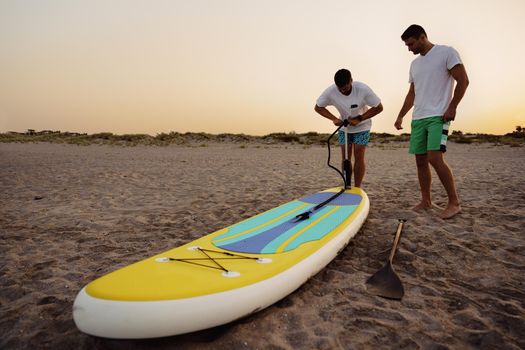 Young man inflating paddle sup board on the sandy beach at sunrise