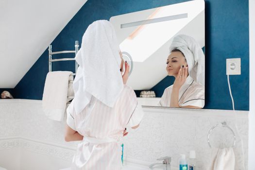 Portrait of cheerful lady in robe and towel on head applying face cream looking at herself in the mirror in the bathroom in the morning. She is smiling while putting cream or lotion on face.