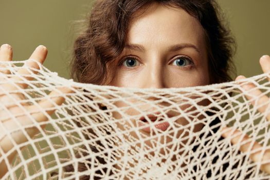 Conceptual Abstract Art. Excited curly beautiful female hold string bag near face seriously looks at camera posing isolated on olive green background. Eco-friendly Natural products concept. Copy space