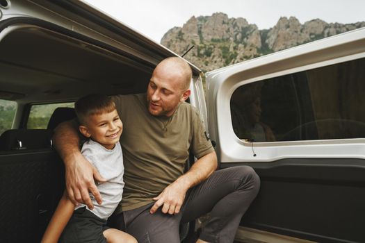 Happy father and son sitting in car trunk on road trip