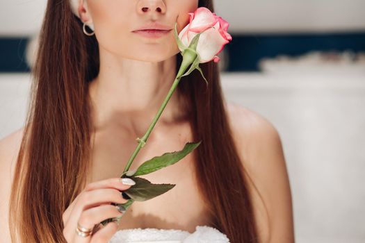 Front view of beautiful tender woman after shower caring about skin in morning. Young long haired lady keeping pink rose, touching face and posing at home. Concept of delicacy and pleasure.