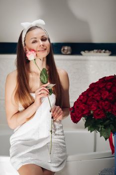 Brunette girl with long hair sitting on bath after morning shower. Pretty lady in white towel looking up and holding flower. Young woman getting bouquet of roses and thinking about boyfriend.