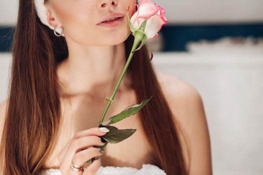 Front view of beautiful tender woman after shower caring about skin in morning. Young long haired lady keeping pink rose, touching face and posing at home. Concept of delicacy and pleasure.