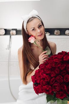 Portrait of gorgeous sensual young brunette woman with headband wrapped in towel sitting in bathroom with bunch of beautiful red roses. She is holding fragile pink rose and smiling at camera. Beauty and skincare concept.