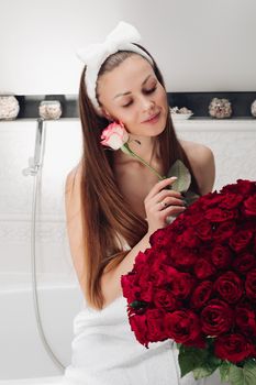 Portrait of gorgeous sensual young brunette woman with headband wrapped in towel sitting in bathroom with bunch of beautiful red roses. She is holding fragile pink rose and smiling at camera. Beauty and skincare concept.