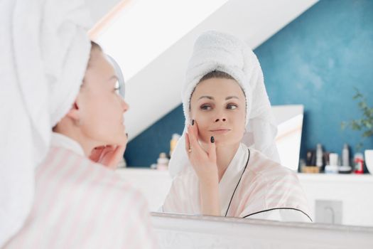 Young woman in white towel and dressing gown looking at camera and applying cream on face in bathroom. Pretty female caring about skin after morning procedures at home. Concept of tenderness.