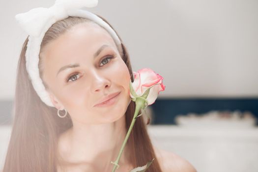 Portrait of sensual beautiful woman in white headband with bow for washing face smiling slightly at camera with fragile pink rose bud at her face.