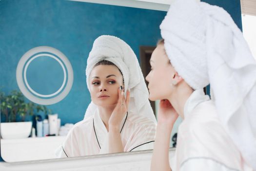 Young woman in white towel and dressing gown looking at camera and applying cream on face in bathroom. Pretty female caring about skin after morning procedures at home. Concept of tenderness.