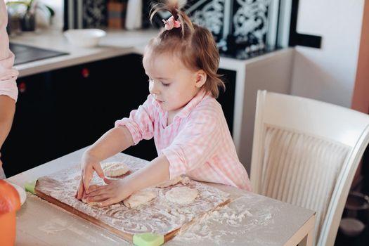 Close up of little serious kid sitting on white chair in pink pajama. Lovely daughter helping her mother preparing breakfast for family. Cute child in flour cooking together with parent at kitchen.