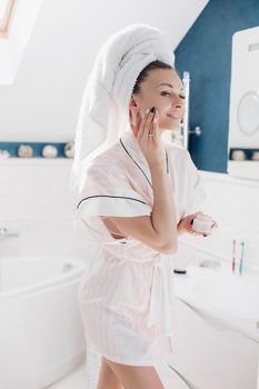 Portrait of cheerful lady in robe and towel on head applying face cream looking at herself in the mirror in the bathroom in the morning. She is smiling while putting cream or lotion on face.