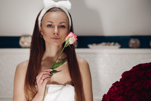 Portrait of sensual beautiful woman in white headband with bow for washing face smiling slightly at camera with fragile pink rose bud at her face.