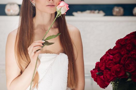 Front view of beautiful tender woman after shower caring about skin in morning. Young long haired lady keeping pink rose, touching face and posing at home. Concept of delicacy and pleasure.